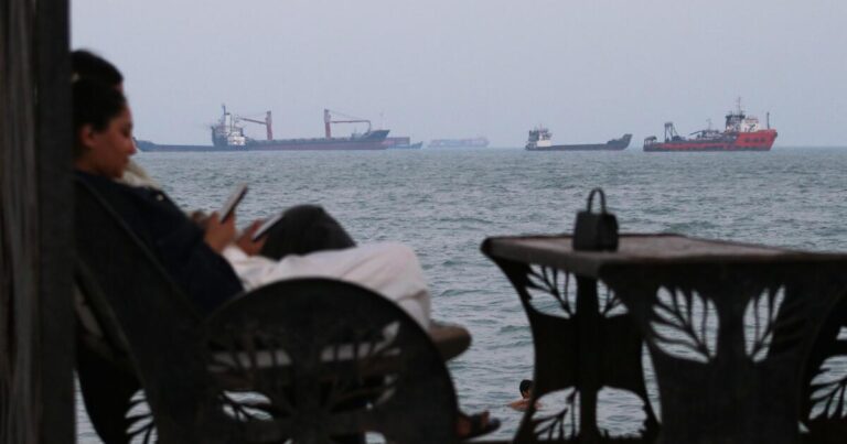 Residents check their cellphones as they sit at a cafe overlooking commercial vessels anchored in the Strait of Hormuz, off