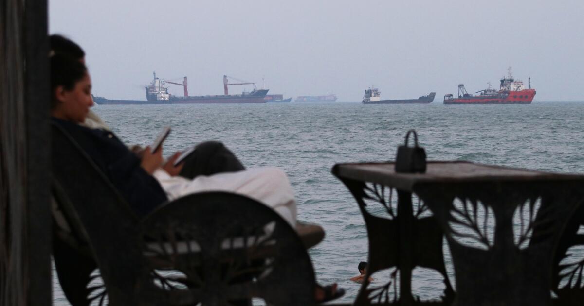 Residents check their cellphones as they sit at a cafe overlooking commercial vessels anchored in the Strait of Hormuz, off