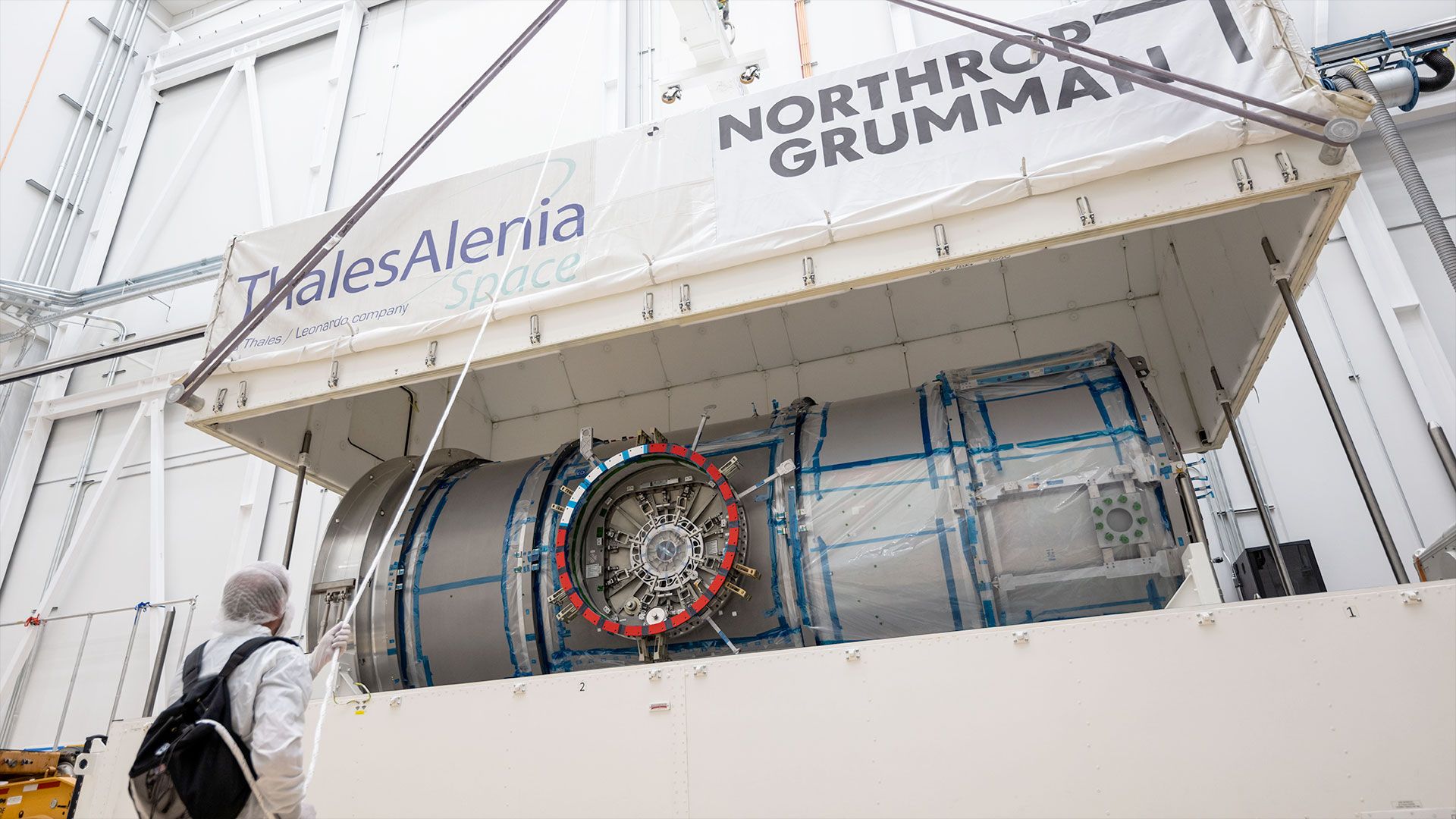 a white top half of a large shipping container is lifted by crane, exposing a metal cylindrical module inside