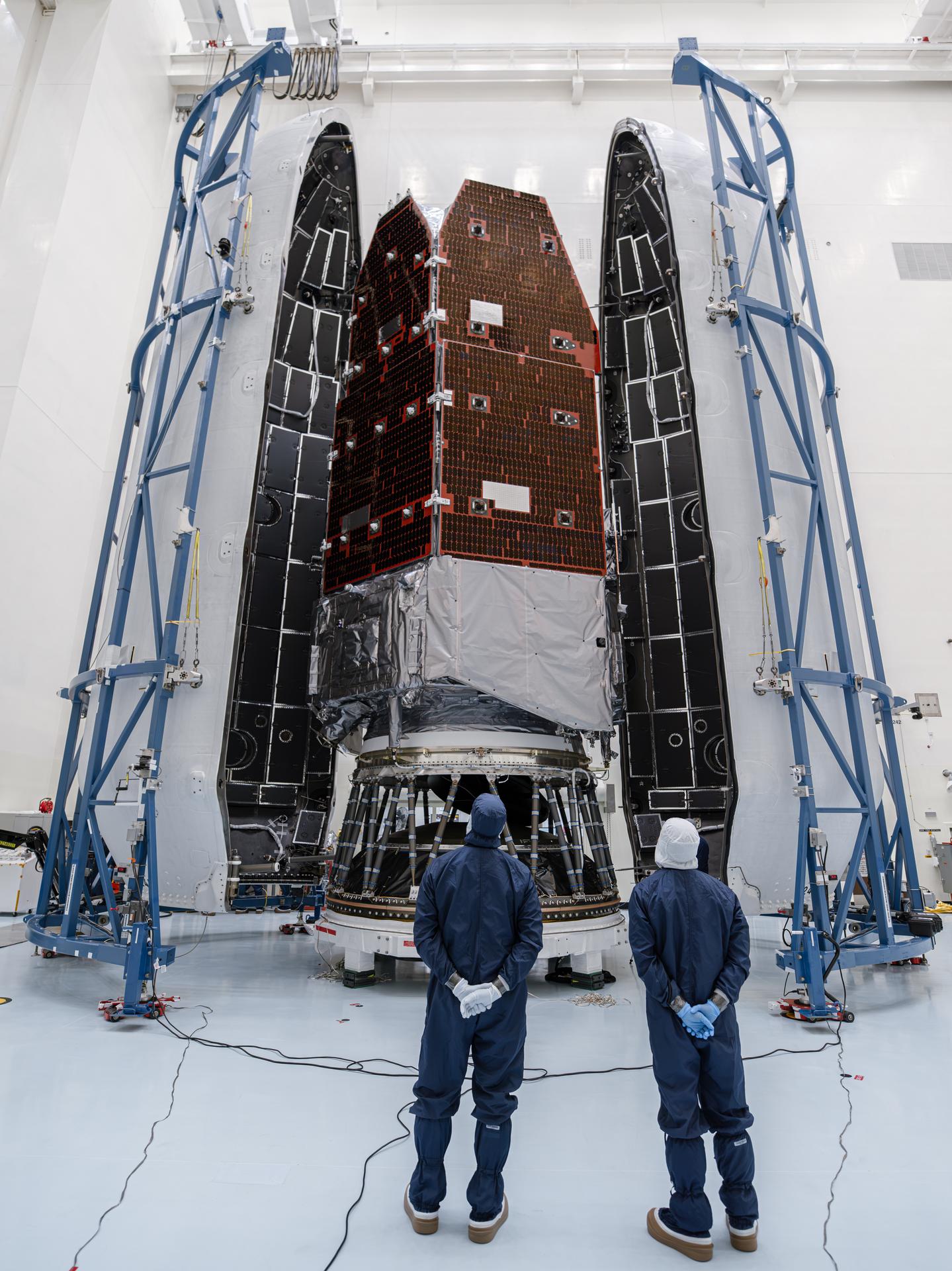 Two technicians in cleanroom suits stand in front of the Roman Space Telescope and payload fairing, which tower above them
