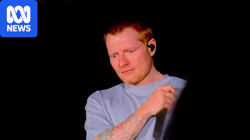 Ed Sheeran wearing a grey shirt looks sad holding a mic at a night-time concert