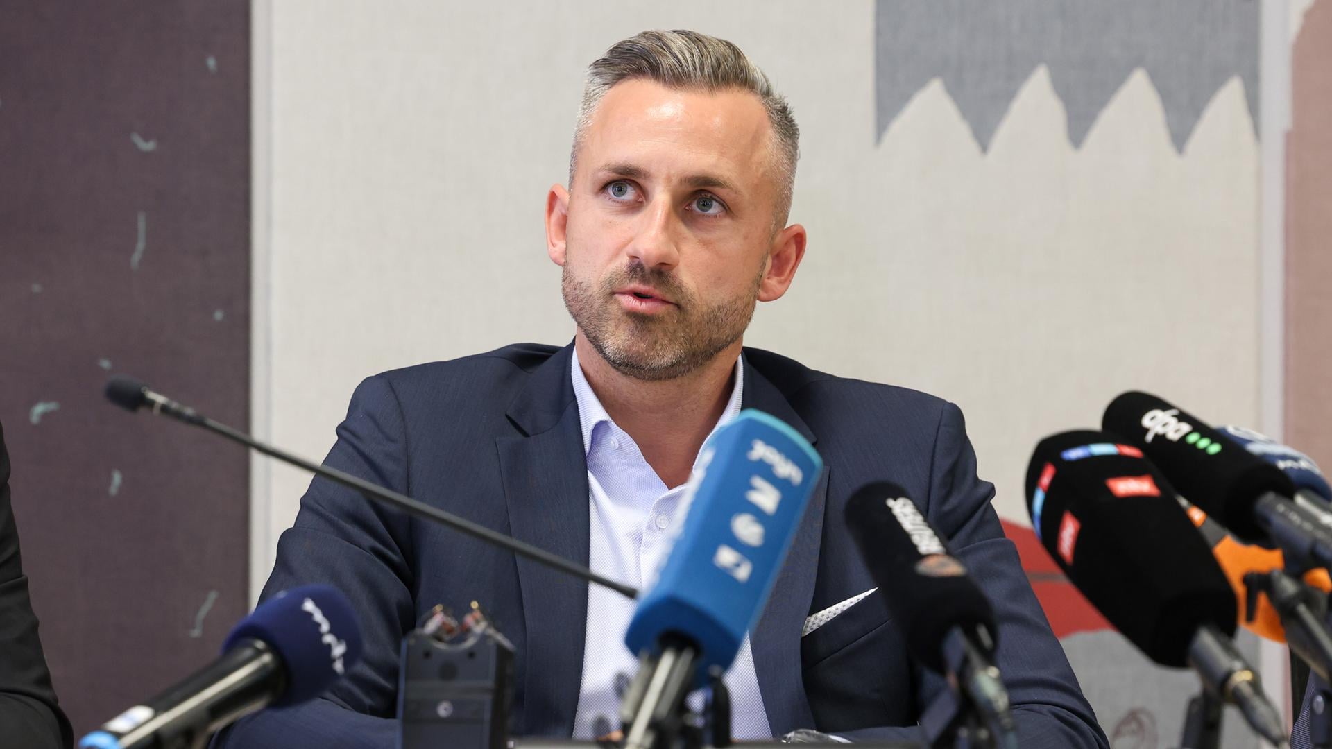 Ulrich Siegmund sitzt an einem Tisch vor mehreren Mikrofonen. Er trägt ein dunkles Jackett und ein hellblaues Hemd