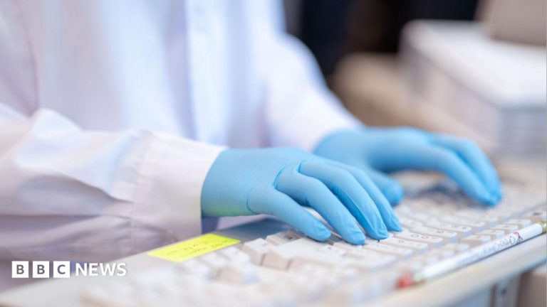 A close-up stock image of a lab worker, wearing a white coat and blue gloves, typing on a keyboard